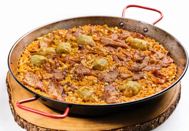 Arroz al horno con trozos de carne y alcachofas, servido en paellera sobre tabla de madera.