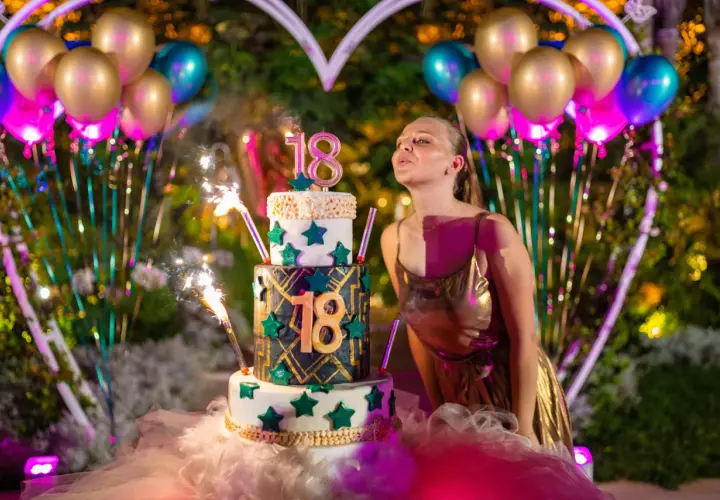 Joven soplando velas en un pastel de tres pisos para celebrar su 18 cumpleaños, rodeada de globos y luces.