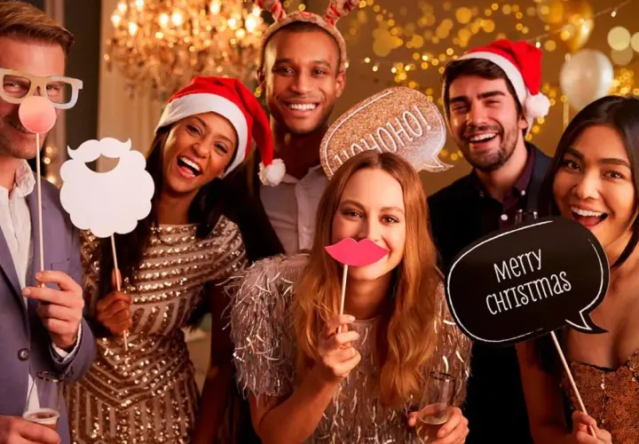Grupo de amigos sonriendo y riendo en un photocall navideño casero, con accesorios como gafas, barba, labios y carteles de "Ho ho ho" y "Merry Christmas".