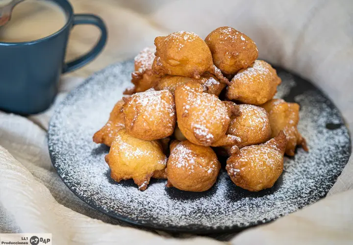 Montón de buñuelos de manzana dorados y espolvoreados con azúcar glas, junto a una taza de café.