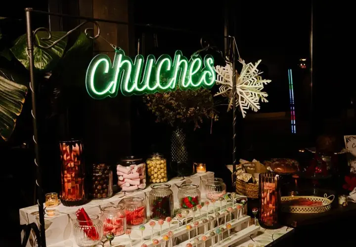 Un letrero de neón verde "Chuchas" ilumina un candy bar lleno de frascos de dulces, piruletas y golosinas.