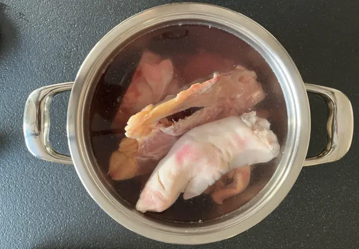 Olla con ingredientes para caldo de huesos: patas de cerdo, huesos y trozos de carne en un líquido oscuro.