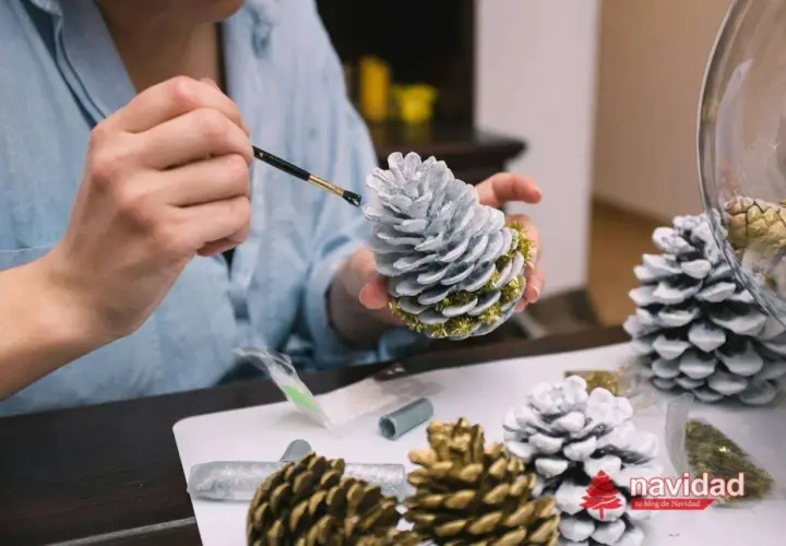 Manos pintando piñas para navidad con pintura plateada y detalles dorados. Decoración festiva en proceso.