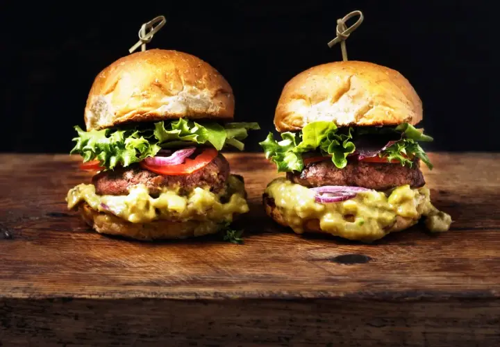 Dos deliciosas hamburguesas caseras con aguacate cremoso, lechuga fresca, tomate y cebolla morada, servidas en panecillos dorados sobre una tabla de madera rústica.