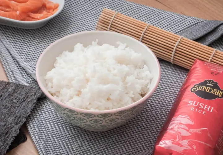 Bol con arroz para sushi, al lado hay salmón, alga nori y un esterilla de bambú.