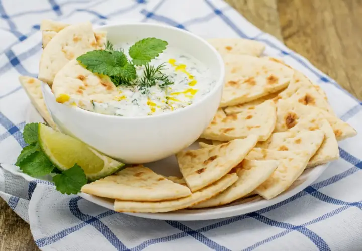 Un cuenco de cremosa salsa tzatziki, decorado con hierbas frescas y un chorrito de aceite, servido con pan de pita crujiente y rodajas de lima.