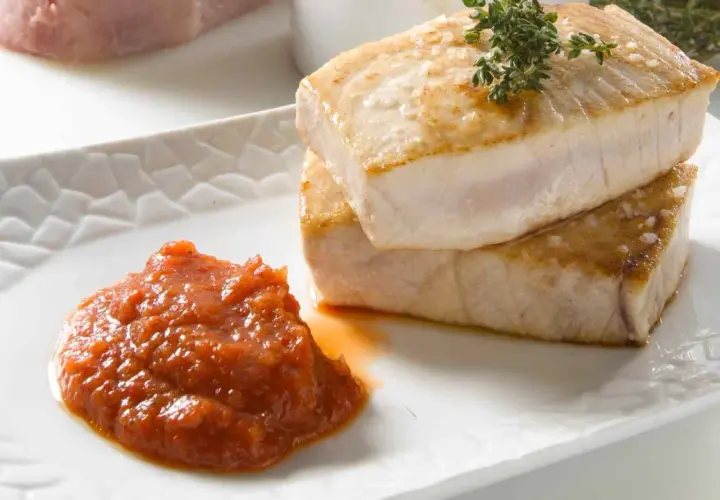 Dos trozos de atún a la plancha, dorados y jugosos, apilados sobre un plato blanco con una porción de salsa de tomate y una ramita de tomillo.
