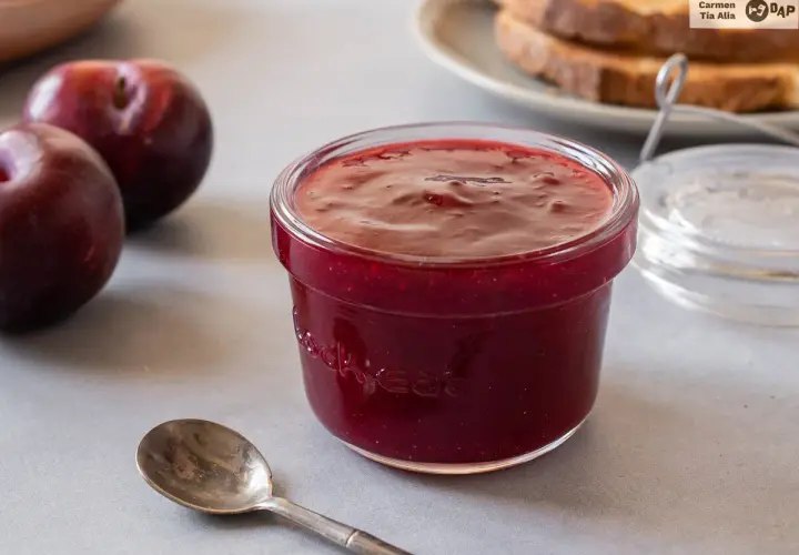 Un tarro de mermelada de ciruela casera, con ciruelas frescas y tostadas al fondo. ¡Perfecta para el desayuno!