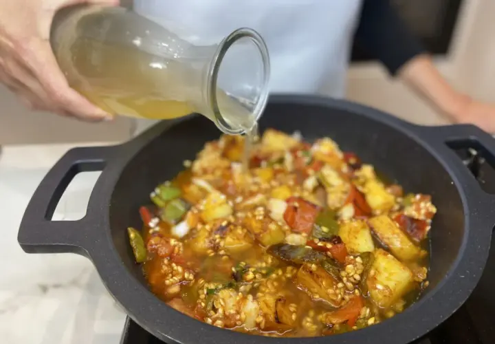 Se vierte caldo sobre un sofrito para paella con pimientos, tomate y patatas en una paellera.