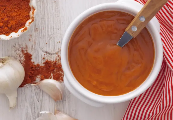 Un cuenco blanco con salsa brava, pimentón en polvo, ajos y una servilleta a rayas rojas.