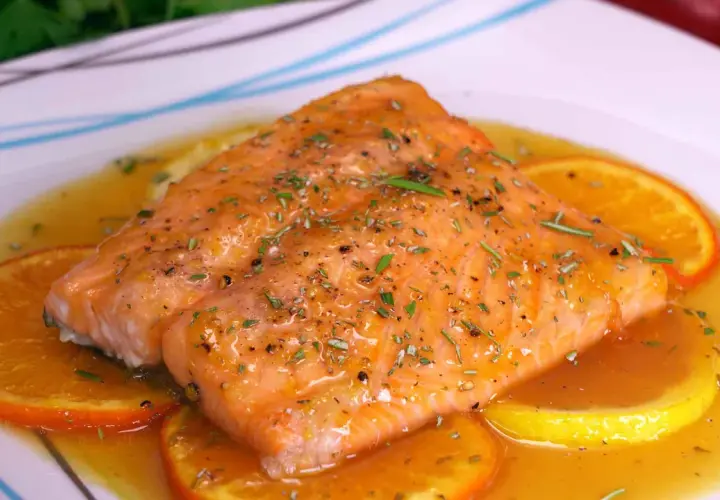 Filete de salmón a la naranja, glaseado y con hierbas, servido sobre rodajas de naranja y limón en una salsa dorada.
