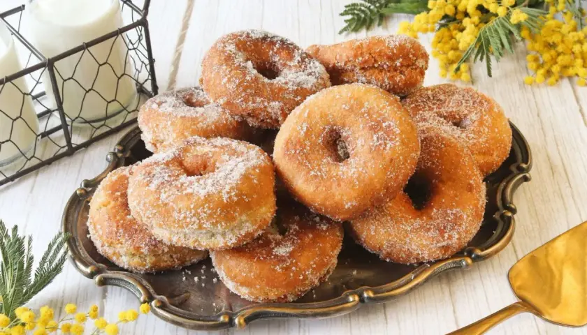 Un plato de rosquillas caseras de toda la vida, espolvoreadas con azúcar, junto a botellas de leche y flores amarillas.