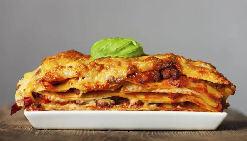 Una porción generosa de lasaña de carne, con capas de pasta, salsa y queso dorado, coronada con una hoja de albahaca fresca.
