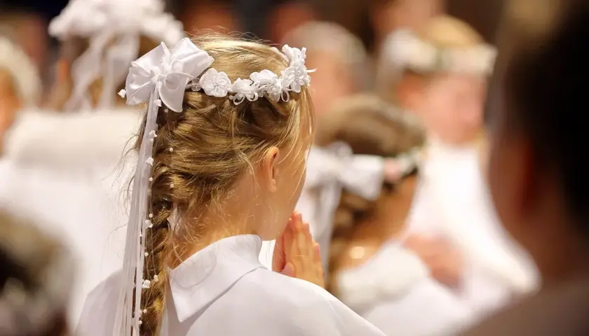Niña con corona de flores y trenza adornada, rezando en una ceremonia de comunión.