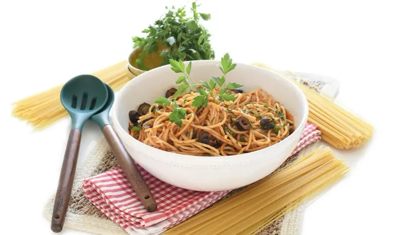Un tazón de espagueti con salsa putanesca, aceitunas negras y perejil fresco, acompañado de pasta cruda y utensilios de cocina.