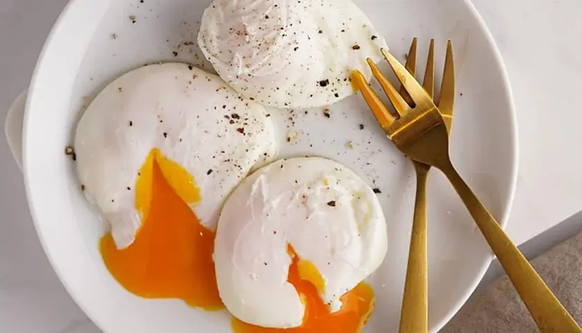 Tres huevos poche perfectamente cocidos, con la yema líquida y dorada, espolvoreados con pimienta negra, listos para disfrutar.