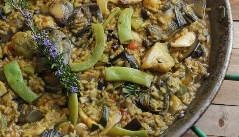 Paella de verduras recién hecha, rebosante de alcachofas, calabacín, pimientos y coliflor, con una ramita de romero y flores moradas.