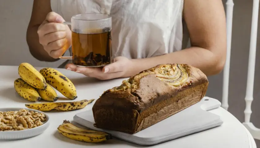 Un delicioso banana bread recién horneado, acompañado de plátanos maduros, nueces y una taza de té humeante.