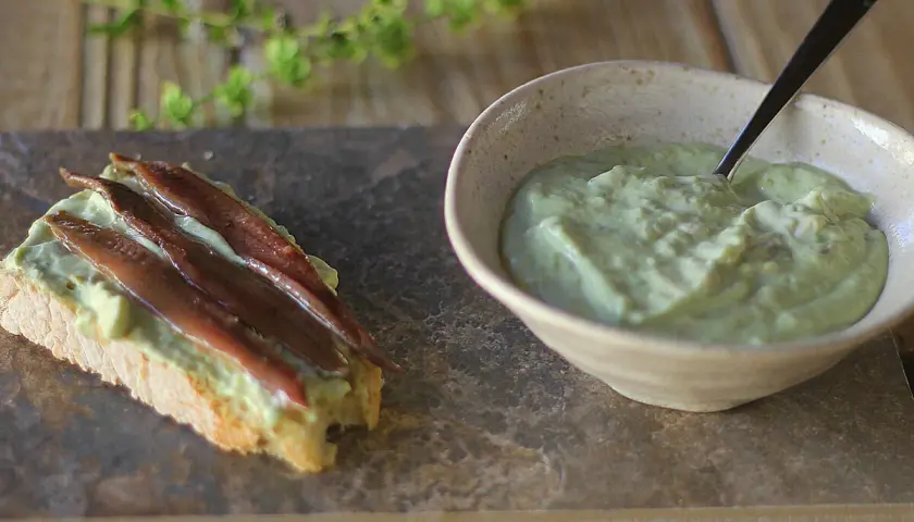 Tosta con mayonesa de aguacate y anchoas. Un cuenco con la cremosa mayonesa de aguacate y una tostada decorada con ella y filetes de anchoa.