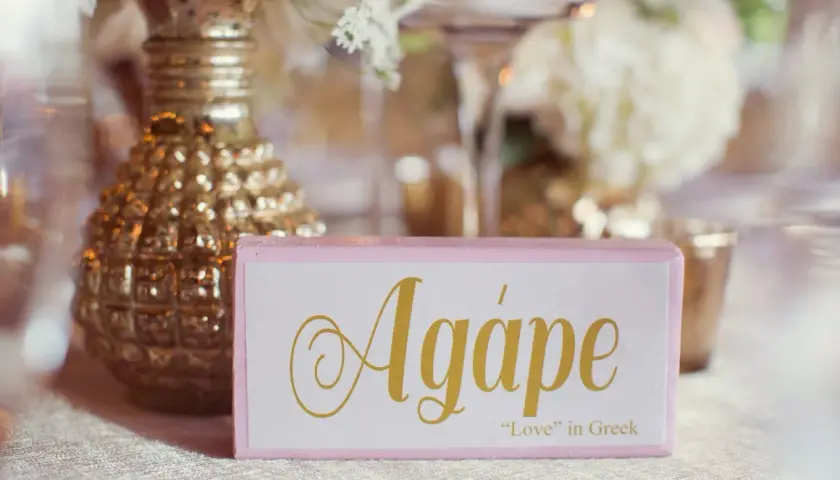 Cartel rosa con la palabra "Agápe" en dorado, junto a un jarrón dorado y flores.
