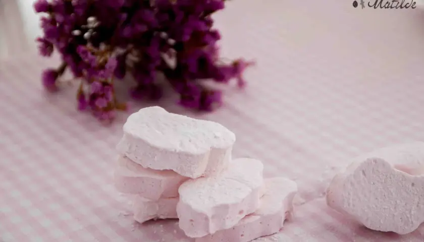 Delicados nubes dulces rosados, espolvoreados con azúcar glas, junto a un ramo de flores moradas sobre un mantel de cuadros.