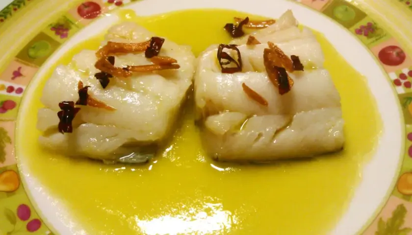 Dos trozos de bacalao al pil pil, cubiertos con aceite de oliva y adornados con ajo frito y guindilla.
