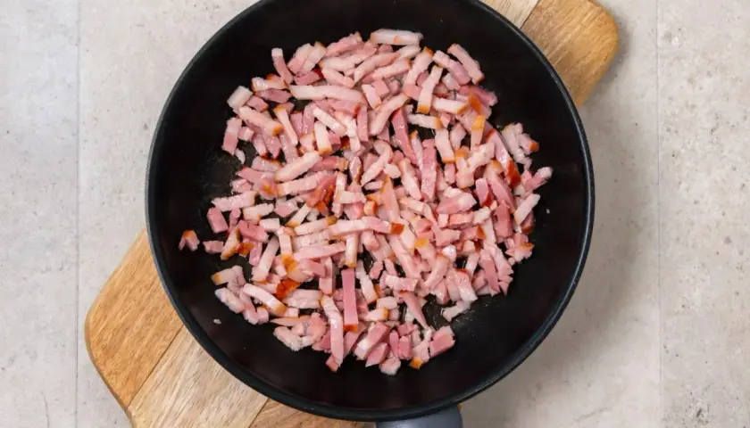 Trozos de bacon crudo en una sartén negra sobre una tabla de madera. Preparando la base para una deliciosa mermelada de bacon.