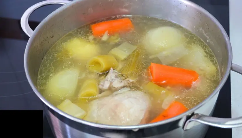 Olla con caldo de pollo casero burbujeante, con trozos de zanahoria, puerro y patata.