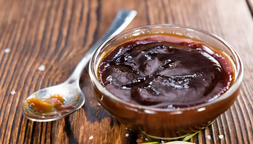 Un cuenco de cristal lleno de deliciosa salsa barbacoa casera, con una cuchara al lado.