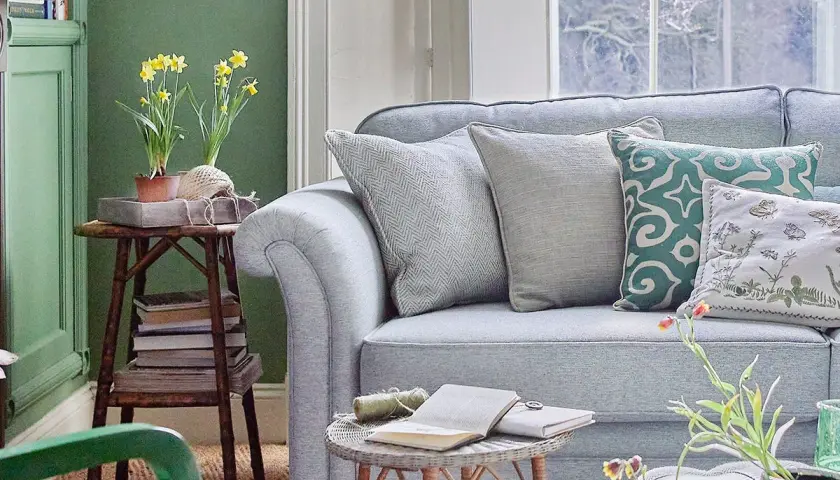 Sofá azul claro con cojines decorativos. Una mesa auxiliar con narcisos y libros complementa la decoración color pastel.
