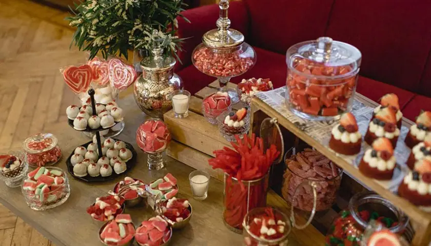 Ideas para como hacer un candy bar para boda: mesa llena de dulces rojos y blancos, pasteles, caramelos y piruletas en forma de corazón.