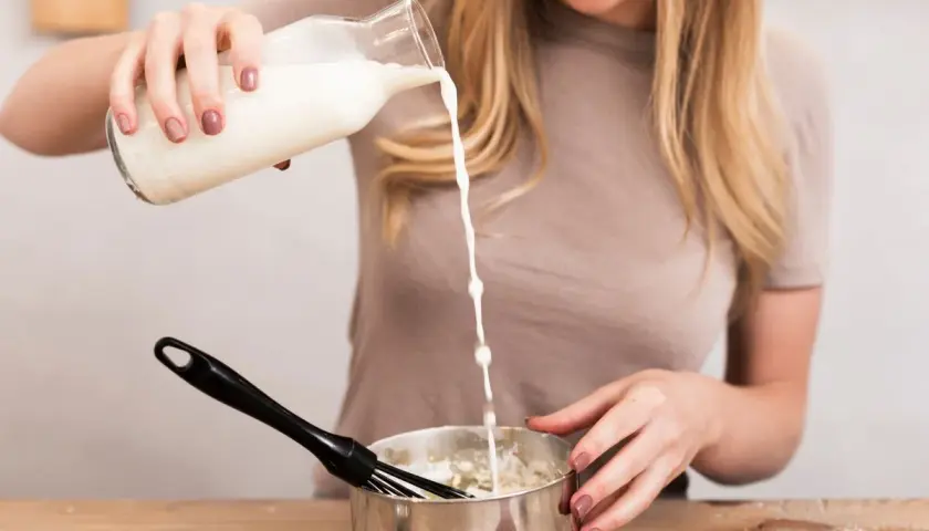 Mujer rubia sonríe mientras vierte leche de una botella a un bol con batidor, preparando algo delicioso.