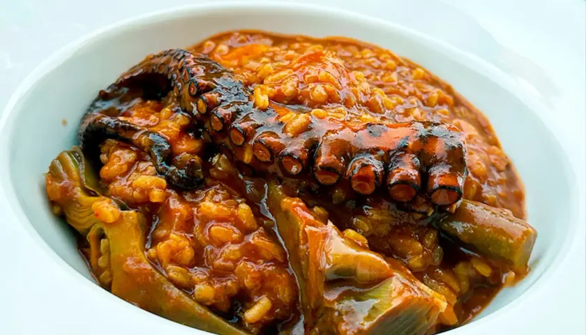 Un tentáculo de pulpo asado reposa sobre un arroz meloso y sabroso, con trozos de alcachofa. Un delicioso arroz con pulpo.