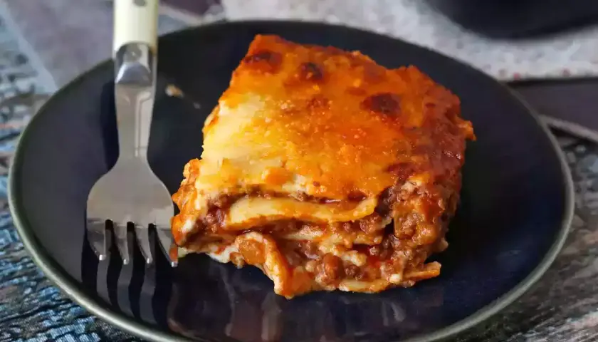 Un trozo de lasaña boloñesa, con capas de pasta, carne y salsa, servido en un plato azul con un tenedor.