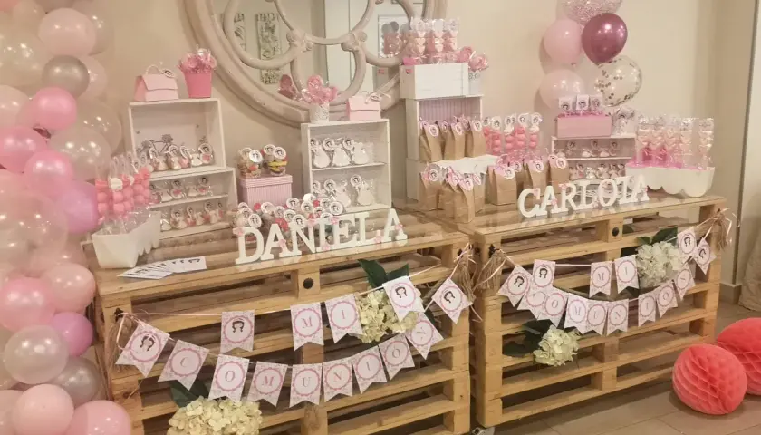 Mesa dulce para comunión de niña, con candy bar casero, globos rosas y blancos, y detalles personalizados.