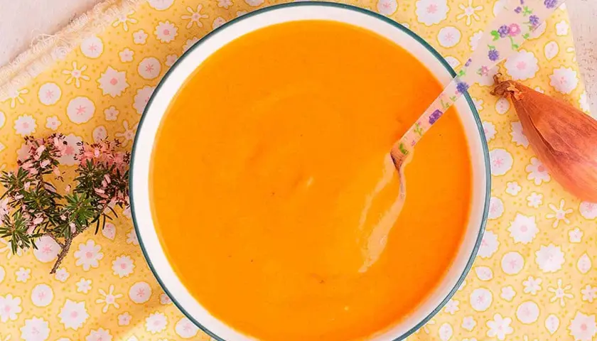 Un tazón de cremosa salsa americana, lista para disfrutar. Una cuchara con flores y una chalota completan la escena.