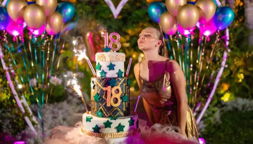 Joven soplando velas en un pastel de tres pisos para celebrar su 18 cumpleaños, rodeada de globos y luces.