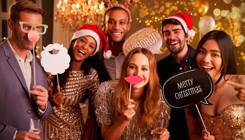 Grupo de amigos sonriendo y riendo en un photocall navideño casero, con accesorios como gafas, barba, labios y carteles de "Ho ho ho" y "Merry Christmas".