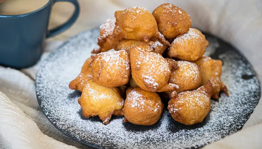 Montón de buñuelos de manzana dorados y espolvoreados con azúcar glas, junto a una taza de café.