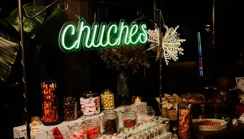 Un letrero de neón verde "Chuchas" ilumina un candy bar lleno de frascos de dulces, piruletas y golosinas.
