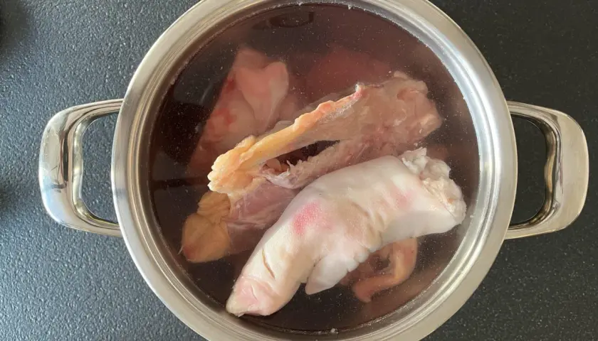 Olla con ingredientes para caldo de huesos: patas de cerdo, huesos y trozos de carne en un líquido oscuro.