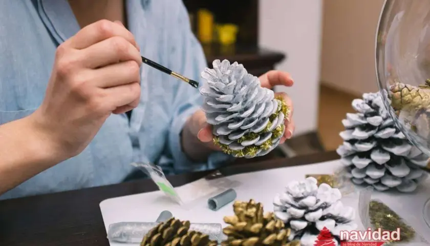 Manos pintando piñas para navidad con pintura plateada y detalles dorados. Decoración festiva en proceso.