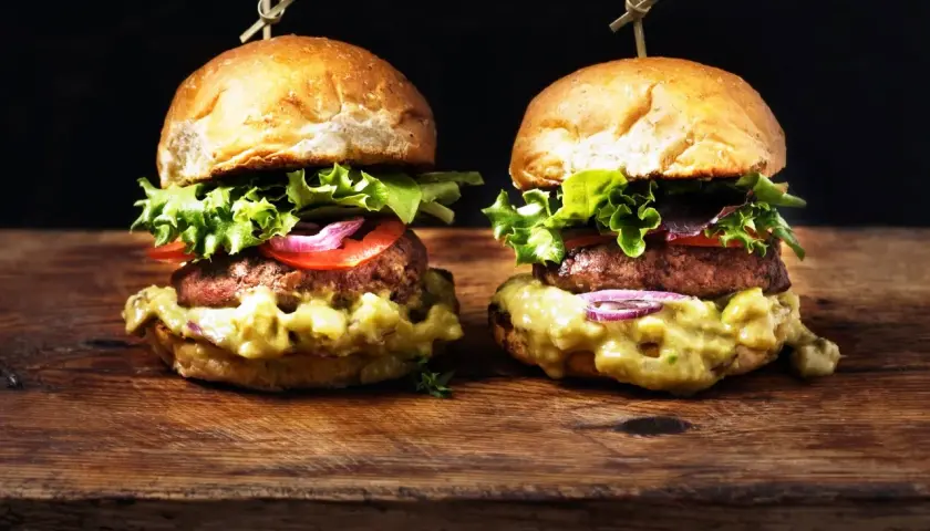 Dos deliciosas hamburguesas caseras con aguacate cremoso, lechuga fresca, tomate y cebolla morada, servidas en panecillos dorados sobre una tabla de madera rústica.