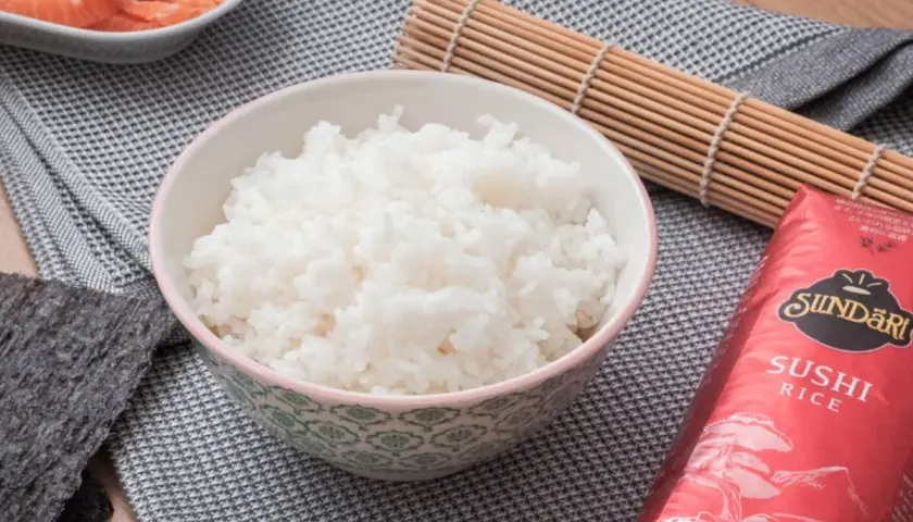 Bol con arroz para sushi, al lado hay salmón, alga nori y un esterilla de bambú.