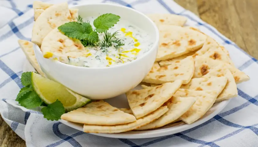 Un cuenco de cremosa salsa tzatziki, decorado con hierbas frescas y un chorrito de aceite, servido con pan de pita crujiente y rodajas de lima.