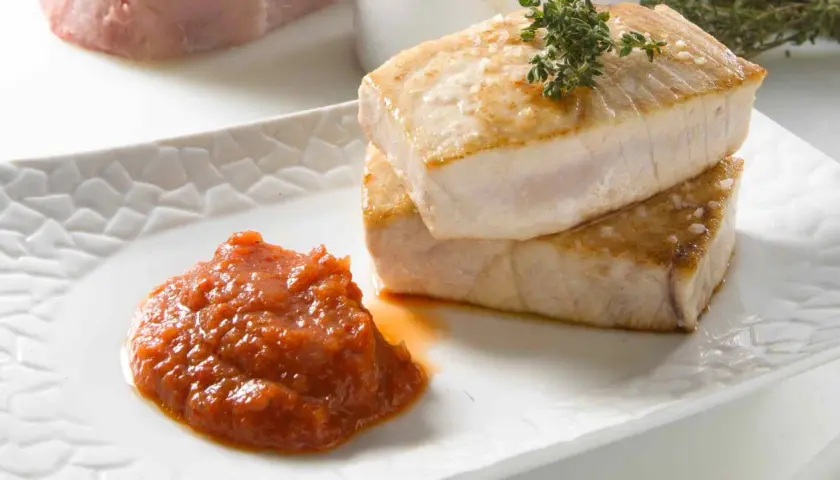 Dos trozos de atún a la plancha, dorados y jugosos, apilados sobre un plato blanco con una porción de salsa de tomate y una ramita de tomillo.