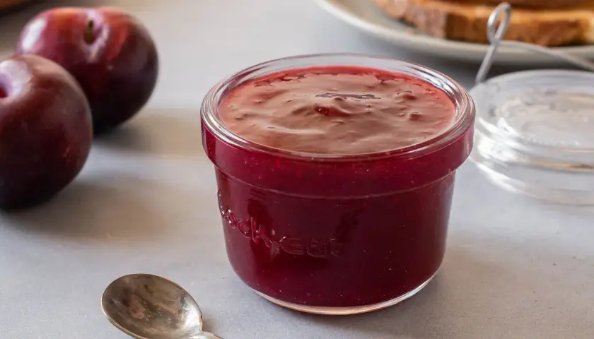 Un tarro de mermelada de ciruela casera, con ciruelas frescas y tostadas al fondo. ¡Perfecta para el desayuno!