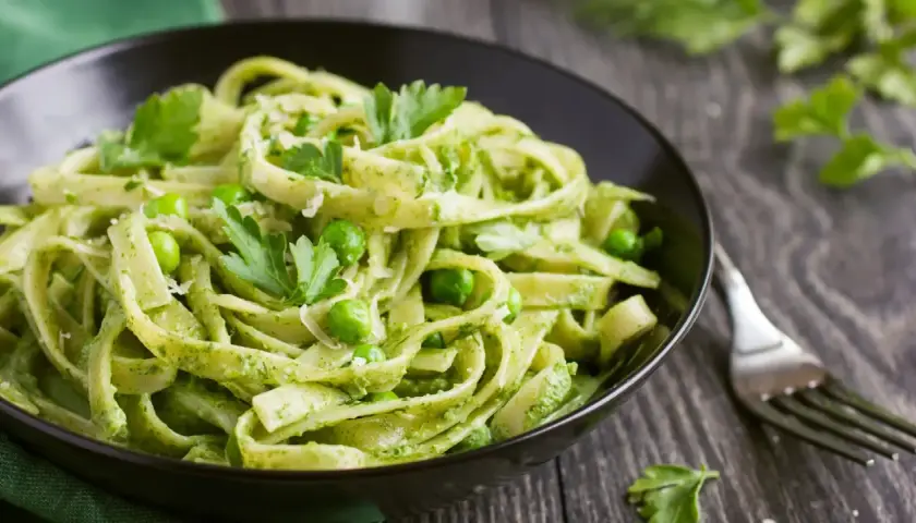 Pasta con espinacas y guisantes, cubierta con perejil fresco y queso rallado, servida en un tazón negro.