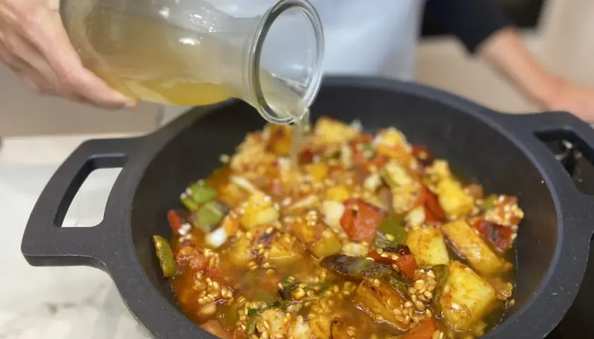 Se vierte caldo sobre un sofrito para paella con pimientos, tomate y patatas en una paellera.