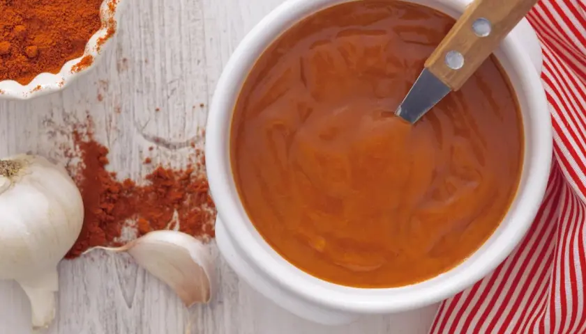 Un cuenco blanco con salsa brava, pimentón en polvo, ajos y una servilleta a rayas rojas.